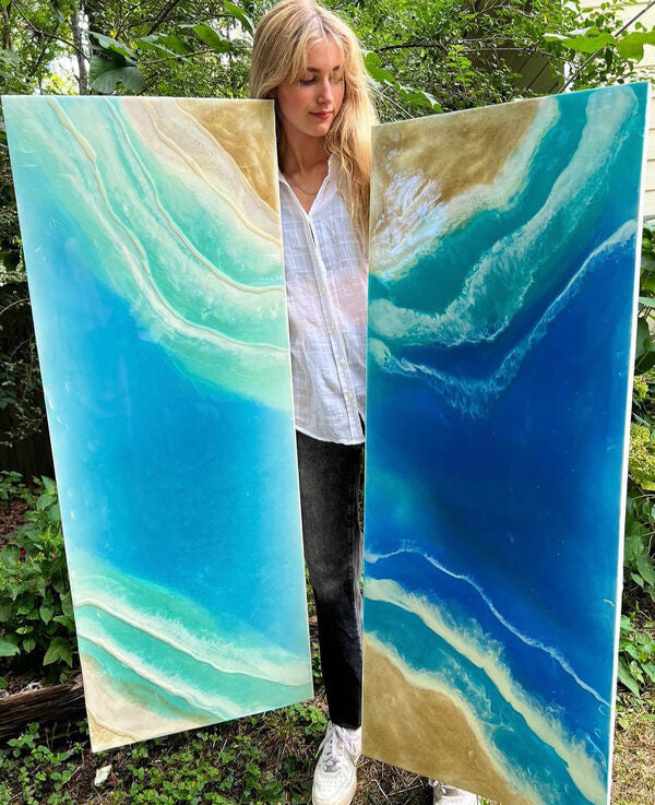 A woman holding up two resin paintings made using epoxy resin and mica pigment powder.